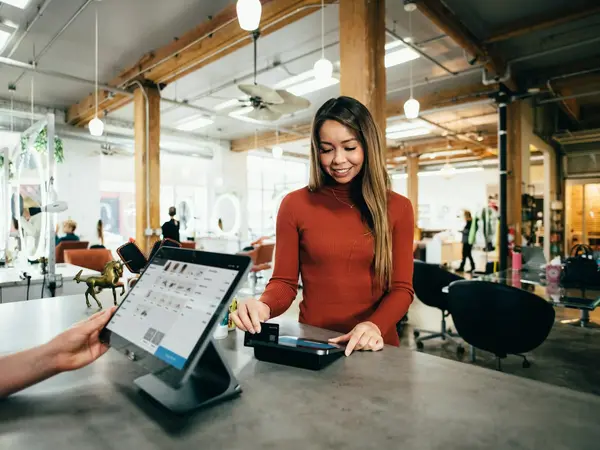 A shop assistant serving a customer at a point-of-sale tablet, representing retail and small businesses.